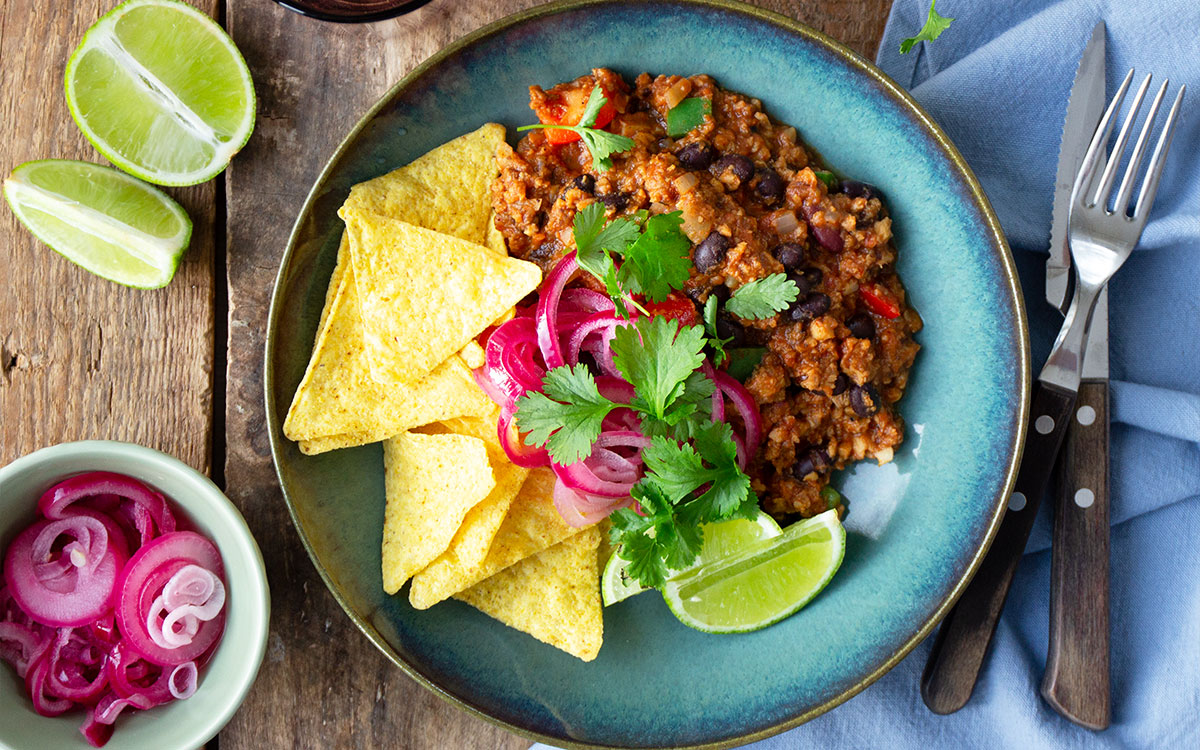 En tallrik med chili sin carne gjord på Baljväxtfärs toppad med picklad rödlök, nachochips och färsk koriander.